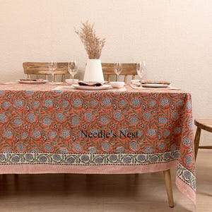 Könnte beinhalten: Ein rechteckiges, orangefarbenes Tischtuch mit floralem Muster auf einem Holztisch. Das Tischtuch zeigt ein sich wiederholendes Design aus weißen und blauen floralen Elementen. Der Tisch ist mit Tellern, Gläsern und einer weißen Vase mit Trockenblumen gedeckt. Der Text "Needle's Nest" ist sichtbar.