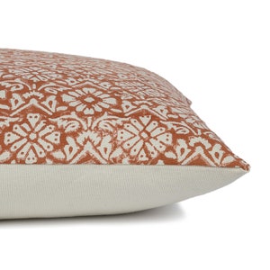 May include: A rust-colored throw pillow with a white geometric pattern. The pillow has a white border and a white back.