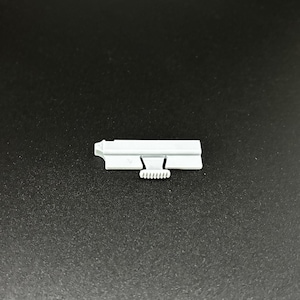 Könnte beinhalten: Eine kleine, hellgraue Metallkomponente mit einem rechteckigen Körper und einem gekerbten, gebogenen Abschnitt. Die Komponente hat ein kleines, zahnradähnliches Merkmal. Das Objekt ist vor einem dunklen Hintergrund platziert.