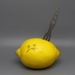 May include: A yellow lemon with a sad face drawn on it, pierced by a black metal fork.