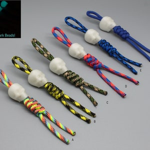 Könnte beinhalten: Sechs Schädel-förmige, im Dunkeln leuchtende Perlen, die an unterschiedlich farbigem Paracord befestigt sind. Das Paracord ist oben an der Perle zu einem Knoten gebunden. Die Perlen sind mit A bis F beschriftet.