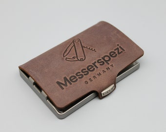 Op de afbeelding: Een bruine leren portemonnee met een metalen frame en een logo met de tekst "Messerspezi Germany" met een zakmesafbeelding.