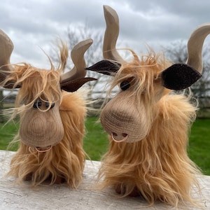 Peut inclure: Deux figurines de vache Highland en bois avec une fourrure brune, des nez noirs et des oreilles en cuir brun. Les vaches ont un anneau autour du nez et sont debout sur une surface en bois blanc.