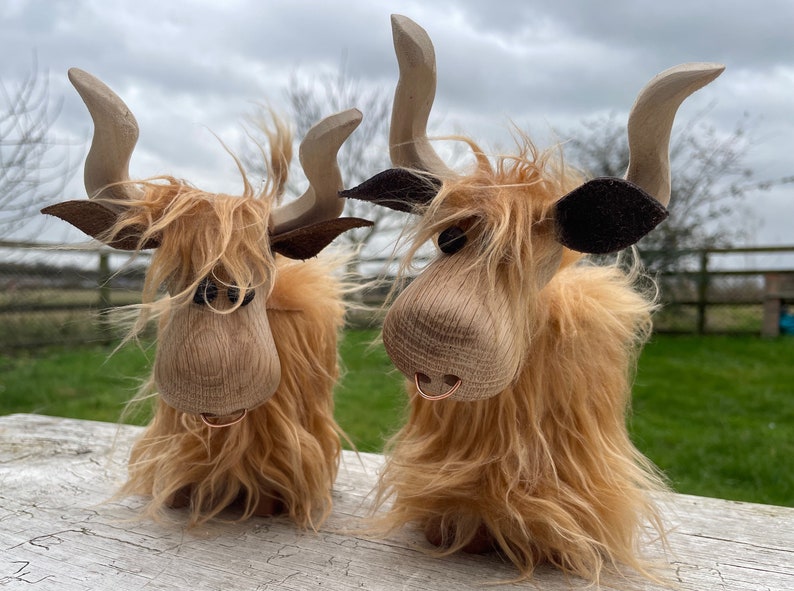 Peut inclure: Deux figurines de vache Highland en bois avec une fourrure brune, des nez noirs et des oreilles en cuir brun. Les vaches ont un anneau autour du nez et sont debout sur une surface en bois blanc.