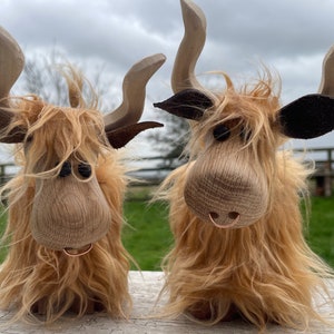 Peut inclure: Deux figurines de vache Highland en bois avec de la fourrure brune, des oreilles noires et un anneau de nez en cuivre. Les vaches sont debout sur une surface en bois.