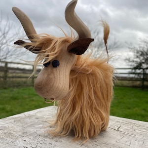 Peut inclure: Une figurine de vache &eacute;cossaise en bois avec un pelage brun, des yeux noirs et un nez en cuir brun. La vache est debout sur une surface en bois.