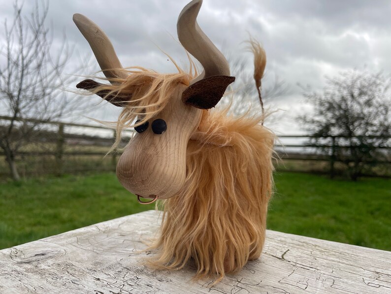 Peut inclure: Une figurine de vache &eacute;cossaise en bois avec un pelage brun, des yeux noirs et un nez en cuir brun. La vache est debout sur une surface en bois.