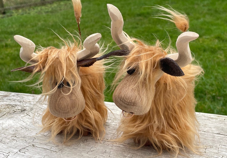 Peut inclure: Deux figurines de vache Highland en bois avec une fourrure brune et des oreilles noires. Les vaches ont un anneau autour du cou et sont debout sur une surface en bois.