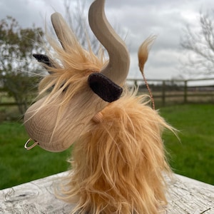 Peut inclure: Une vache Highland en bois jouet avec une longue fourrure brun clair hirsute. La vache a une t&ecirc;te en bois avec un anneau dans le nez et de grandes cornes en bois courb&eacute;es. La vache est debout sur une surface en bois blanc.