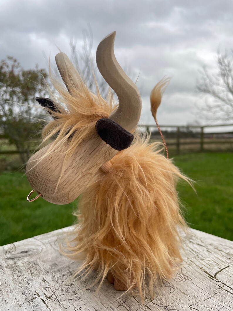 Peut inclure: Une vache Highland en bois jouet avec une longue fourrure brun clair hirsute. La vache a une t&ecirc;te en bois avec un anneau dans le nez et de grandes cornes en bois courb&eacute;es. La vache est debout sur une surface en bois blanc.