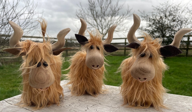 Peut inclure: Trois figurines de vache &eacute;cossaise en bois avec une fourrure brune, des yeux noirs et des cornes en bois. Les vaches sont debout sur une surface en bois.