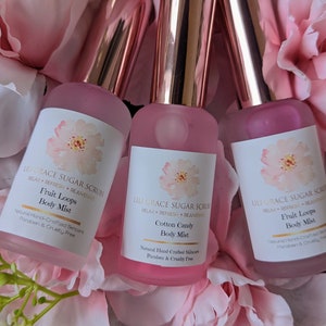 May include: Three pink glass bottles with gold caps and white labels. The labels have a floral design and text that reads "Lil Grace Sugar Scrubs Relax Refresh Rejuvenate" and "Fruit Loops Body Mist" or "Cotton Candy Body Mist". The bottles are on a pink and white floral background.