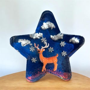 Könnte beinhalten: Sternförmige Harzdekoration mit Winterthema. Das blaue Harz enthält eine Hirschfigur, silberne Schneeflocken und flauschige weiße Wolken. Die Basis des Sterns hat rosa Glitzer. Eine festliche Weihnachtsdekoration.