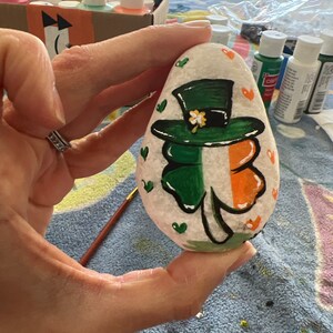 May include: A painted rock featuring a green shamrock and a leprechaun's hat. The shamrock is outlined in black and has a white flower in the centre. The hat is green with a black band and a white buckle. The rock is surrounded by small white hearts.
