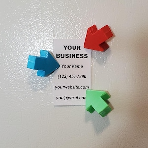 Pode incluir: Um cartão de visita com o texto "YOUR BUSINESS", "Your Name", um número de telefone, um site e um endereço de e-mail. Três ímãs em forma de seta azuis, vermelhos e verdes circundam o cartão. O cartão e os ímãs estão em uma superfície branca.