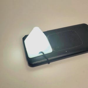 May include: A bright white triangular light is positioned on a black rectangular device. A black cord connects to the light. The device looks like a wireless charger. The light is luminous, brightening the area around it.