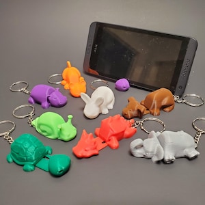 May include: A collection of colorful 3D-printed animal keychains, including a turtle, snail, hippo, rabbit, and elephant. Each keychain is attached to a silver keyring. A black smartphone is propped up in the background.