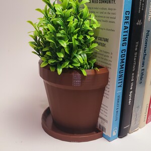 May include: A brown ceramic plant pot with a green artificial plant inside, used as a bookend. The bookend is holding up a book titled "Creating Community" by Stanley & Willits. The book is next to other books, including "The Starfish and the Spider" and "Nicomachean Ethics".