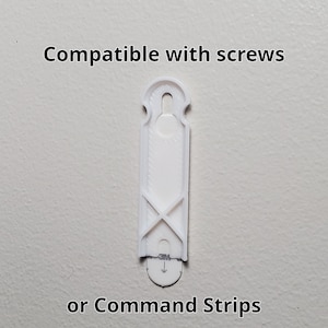 K&ouml;nnte beinhalten: Ein wei&szlig;er Wandhaken aus Kunststoff mit dem Text "Compatible with screws oder Command Strips." Der Haken hat eine rechteckige Form mit einem zentralen Schlitz und einem gew&ouml;lbten Oberteil. Er hat ein X-f&ouml;rmiges Design in der N&auml;he des Bodens.
