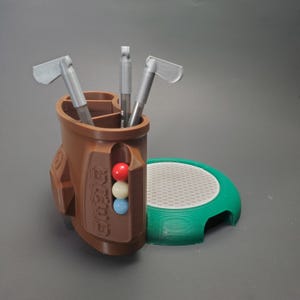 May include: A brown golf bag-shaped desk organizer with three golf clubs and a pen inside. The organizer has a side compartment with three colored golf balls. A green and white golf tee holder is next to the organizer.