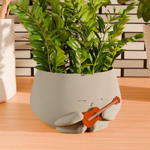 Peut inclure: Un pot en céramique gris en forme de lapin de dessin animé tenant une petite guitare en bois. Le pot est rempli de plantes vertes feuillues.