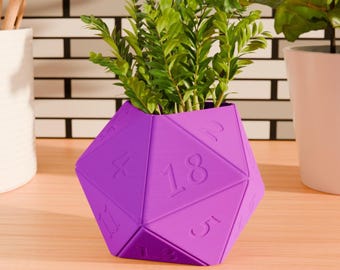 Könnte beinhalten: Lilafarbener D20-Würfel-Pflanztopf mit einer kleinen grünen Pflanze, die darin wächst. Der Topf hat auf jeder Seite die Zahlen von 1 bis 20.