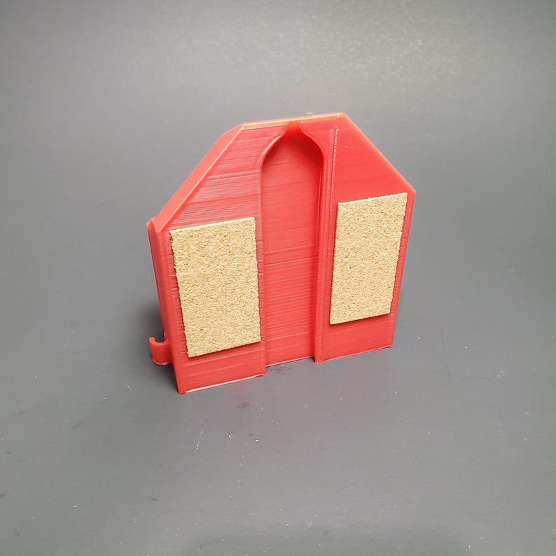 K&ouml;nnte beinhalten: Ein rotes, 3D-gedrucktes Objekt in Hausform. Es hat zwei rechteckige Korkpads und einen bogenf&ouml;rmigen Ausschnitt. Das Objekt ist daf&uuml;r konzipiert, etwas zu halten, m&ouml;glicherweise ein Telefon oder einen kleinen Gegenstand. Das Design ist einfach und funktional.