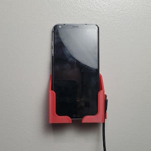 K&ouml;nnte beinhalten: Ein roter, 3D-gedruckter Telefonhalter an einer wei&szlig;en Wand, der ein schwarzes Smartphone h&auml;lt. Ein schwarzes Ladekabel ist an das Telefon angeschlossen. Der Bildschirm des Telefons ist dunkel und spiegelt die Umgebung wider.