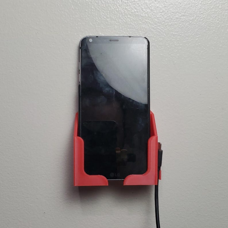 K&ouml;nnte beinhalten: Ein roter, 3D-gedruckter Telefonhalter an einer wei&szlig;en Wand, der ein schwarzes Smartphone h&auml;lt. Ein schwarzes Ladekabel ist an das Telefon angeschlossen. Der Bildschirm des Telefons ist dunkel und spiegelt die Umgebung wider.