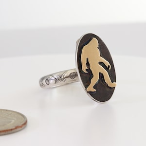 May include: A silver ring with an oval black face featuring a gold-colored Bigfoot silhouette. The ring band has etched designs. A U.S. quarter is in the foreground.
