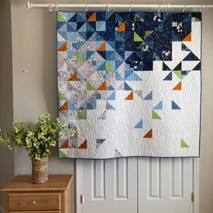 May include: A large, geometric quilt with a blue border is displayed on a wall. The quilt features a pattern of triangles in shades of blue, orange, and green against a white background. The quilt is hanging above a white door.