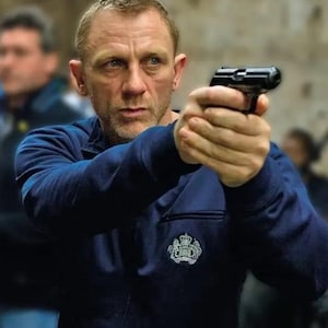 Puede incluir: Un hombre con una chaqueta azul marino con cremallera, sosteniendo una pistola negra, con expresión seria. La chaqueta tiene un emblema blanco en el pecho. El fondo está borroso, sugiriendo un entorno interior.