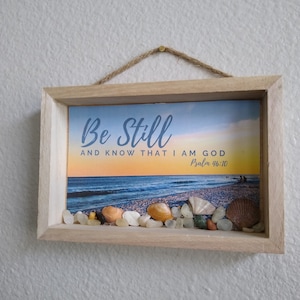 May include: A framed print with a beach scene and the text "Be Still and Know That I Am God, Psalm 46:10". The frame is made of wood and has a rope hanger. There are seashells and small stones in the frame.