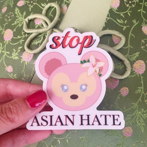 May include: A pink sticker featuring a cartoon bear with a flower and the words "stop ASIAN HATE" in red and black. The sticker is held against a green floral background.