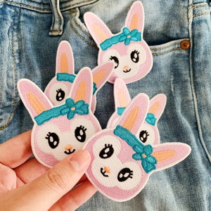 May include: Embroidered patches featuring a pink bunny face with a blue headband and flower. The patches have black eyes, a white face, and pink ears. The patches are on a denim background.