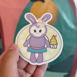 May include: A round sticker featuring a cartoon bunny character in shades of purple, blue, and white. The bunny wears a flower and holds a soft-serve ice cream cone. The background is a light yellow with white polka dots.