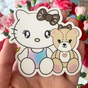 Könnte beinhalten: Ein gestanzter Aufkleber mit einer weißen Cartoon-Katze mit einer braunen Schleife und einem kleinen hellbraunen Teddybären mit einer braunen Schleife und einem rosa Herzanhänger. Die Katze trägt ein blau-violettes Outfit.