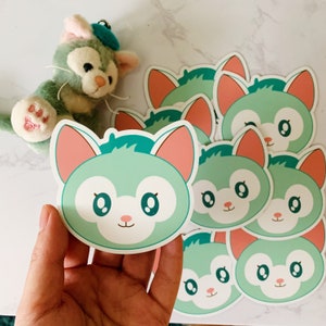 May include: A collection of stickers featuring a cartoon cat face with mint green fur, pink ears, and a small pink nose. The stickers are held in a hand, with a plush cat toy in the background.