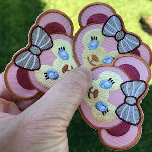 May include: Embroidered patches featuring a bear design with a bow. The patches have a light pink background with a darker pink accent. The bear's face is yellow with blue eyes and a brown nose. The bow is gray and white.