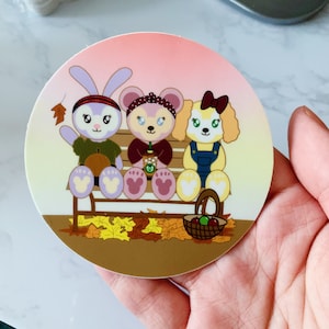 May include: A round sticker featuring three cartoon characters sitting on a bench in autumn. The characters include a bunny, a bear, and a dog. The bench is surrounded by fall leaves and a basket of apples.