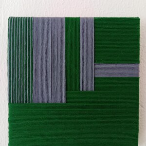 Puede incluir: Una obra de arte textil cuadrada y tejida. La obra presenta un diseño geométrico con rayas verticales de hilo gris y verde sobre un fondo verde. Una franja gris horizontal intersecta el diseño. La pieza está montada sobre una superficie blanca.