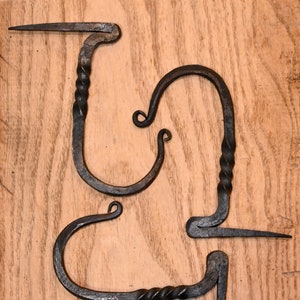 May include: Two black metal hooks with a twisted design. The hooks are made of wrought iron and have a rustic look.