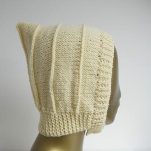 May include: Cream-coloured knitted bonnet with a pointed top. The bonnet features vertical ribbed detailing and a textured knit pattern. The brim and edges have a distinct, tighter knit. The bonnet is displayed on a mannequin head.