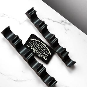 May include: Two black, curved, ridged holders and a black rectangular tag with white text. The holders have a wave-like design with four curved ridges. The tag reads "ENGRAVE. PRINT. CREATE. DIGITAL Timber."