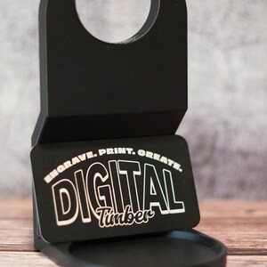 May include: Black plastic drink holder with a round base and a handle. The holder has a small rectangular plate with the text "ENGRAVE. PRINT. CREATE. DIGITAL Timber" in white lettering.