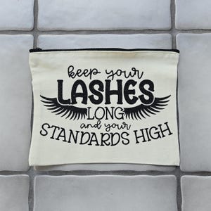 Könnte beinhalten: Eine cremefarbene Kosmetiktasche mit einem schwarzen Reißverschluss. Die Tasche trägt den Text "keep your LASHES LONG and your STANDARDS HIGH" in schwarzer Schrift und ein dekoratives Flügeldesign.