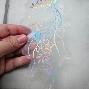 Op de afbeelding: Een transparante, iriserende sticker met een kwalontwerp. De kwal is omlijnd met een holografische afwerking en heeft bloemen eromheen.