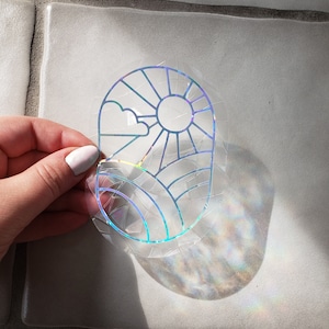 May include: A clear, holographic sticker with a sun, clouds, and landscape design. The sticker has a geometric style with lines and shapes, creating a rainbow effect when light hits it. The sticker is held up by a hand.