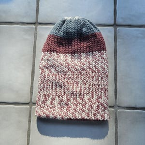 May include: A knitted beanie hat with a color block design. The top section is a muted blue, followed by a burgundy band, and the lower portion is a mix of burgundy and white. The hat is made of a soft, textured yarn.