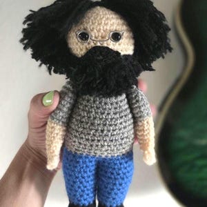 May include: A handmade crochet doll with a black beard and hair, wearing a gray shirt and blue pants. The doll has small wire-rimmed glasses and is approximately 7 inches tall.